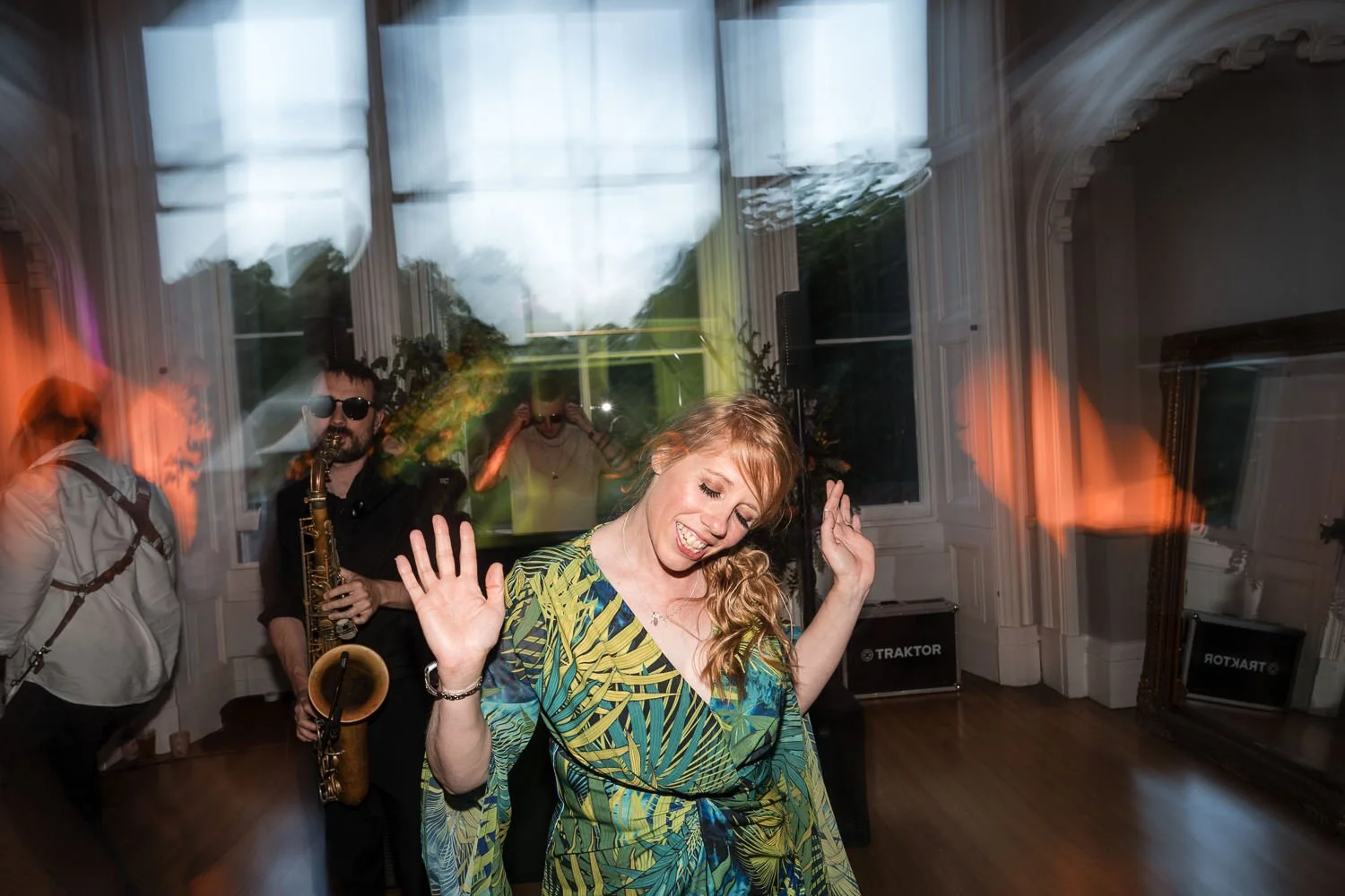 People dancing and enjoying music at a party with a woman in the foreground dancing with a big smile, a saxophone player and a person taking photos in the background, with large windows and decorative interior.
