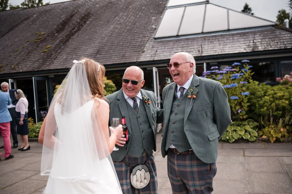 A bride in a white wedding dress and veil converses with two older men in green blazers and plaid kilts at an outdoor wedding reception, smiling and holding drinks, with other guests in the background at Cruin Loch Lomond