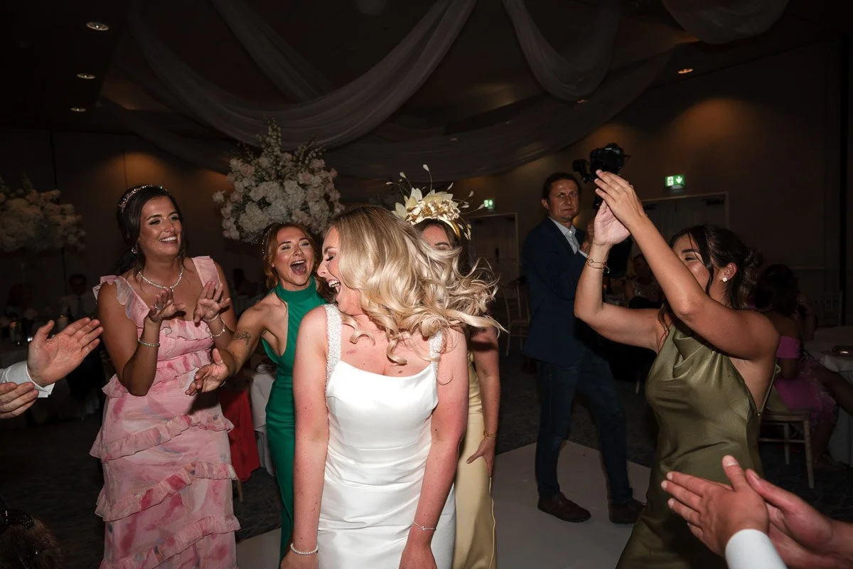 Women dancing and celebrating at a wedding reception, with the bride in a white wedding dress at the centre, surrounded by other women in colourful dresses and a man in a blue suit at Fairmont, St. Andrews