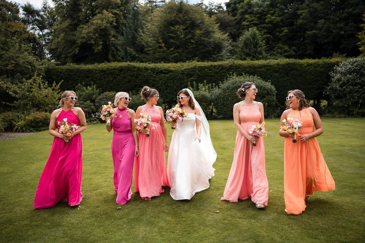 Bridal party of six women in colourful dresses walking on green lawn, smiling and holding bouquets, with trees and bushes in the background at Balbirnie House