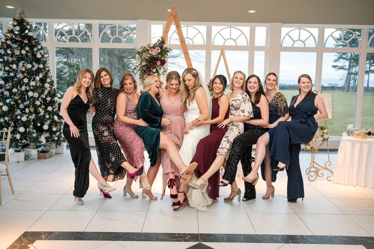 A group of women in formal dresses and high heels, smiling and sitting on a bench, celebrating a wedding, with a Christmas tree and decorated interior in the background at Lochgreen, Troon