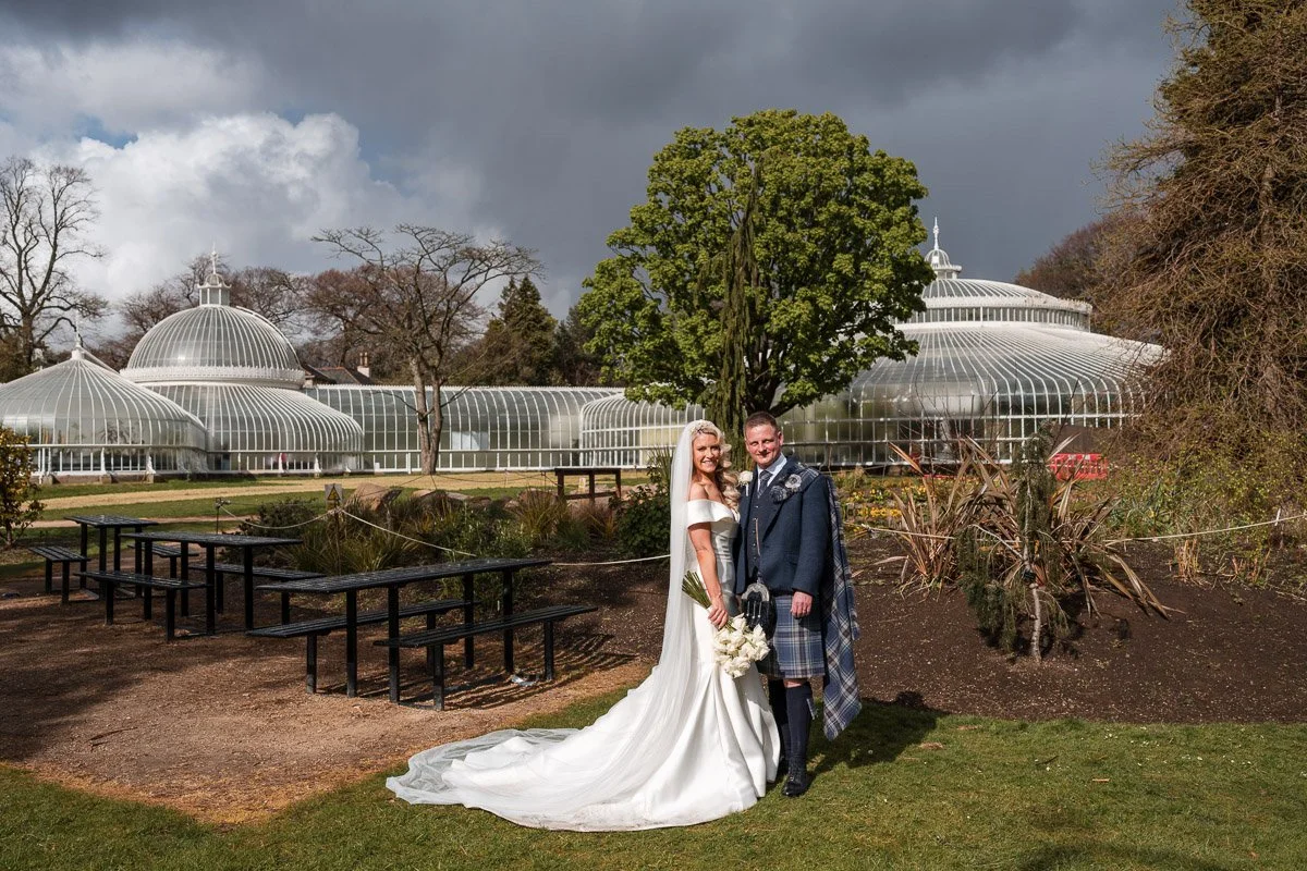 Bride and groom in wedding attire standing outside in front of a large glass greenhouse on a partly cloudy day at Glasgow Botanic Gardens
