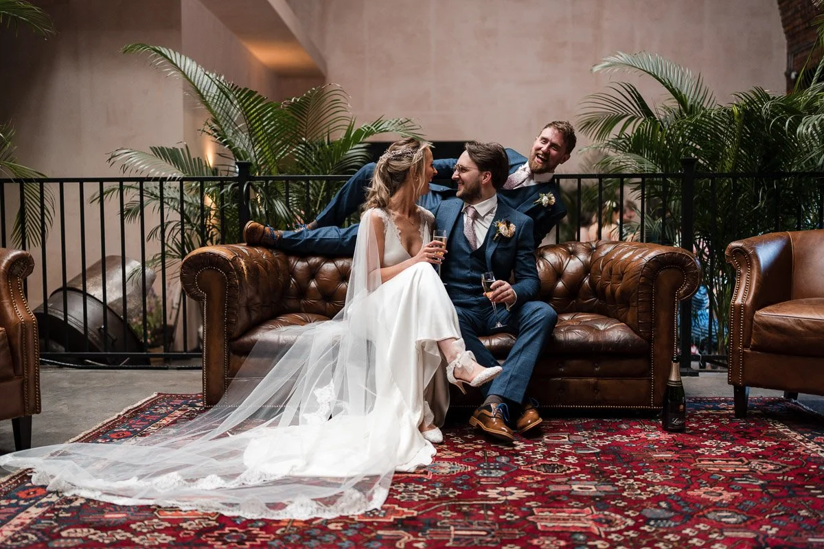 A bride and groom in wedding attire sit on a brown leather sofa, holding champagne glasses, smiling at each other, with a third man in a blue suit leaning over them, all in an indoor setting with plants and a decorative rug.