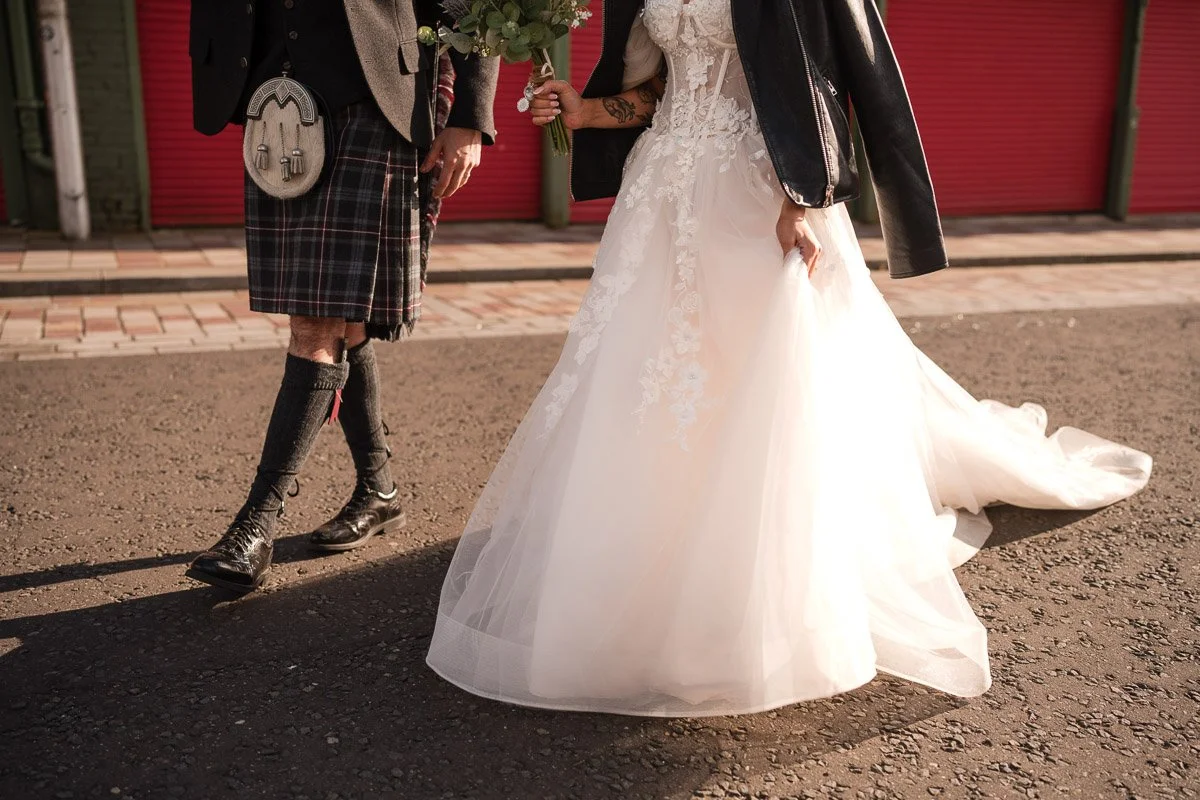 A bride in a white wedding dress holding a bouquet, standing next to a person dressed in a kilt, black shoes, and a leather jacket on a street with red doors in the background at a Barras elopement