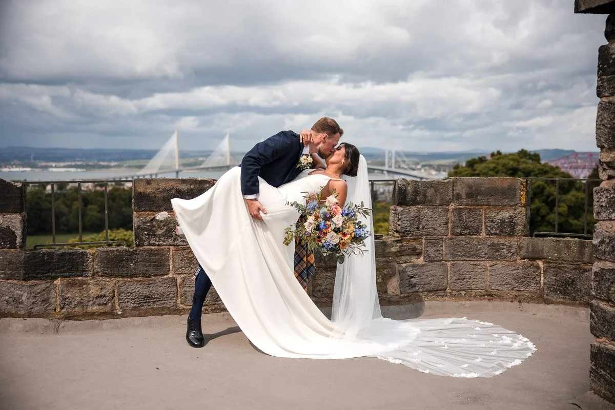 A bride in a white wedding dress holding a colourful bouquet is being dipped by a groom in a kilt while kissing on a stone patio on Dundas Castle roof the Forth bridges behind them.