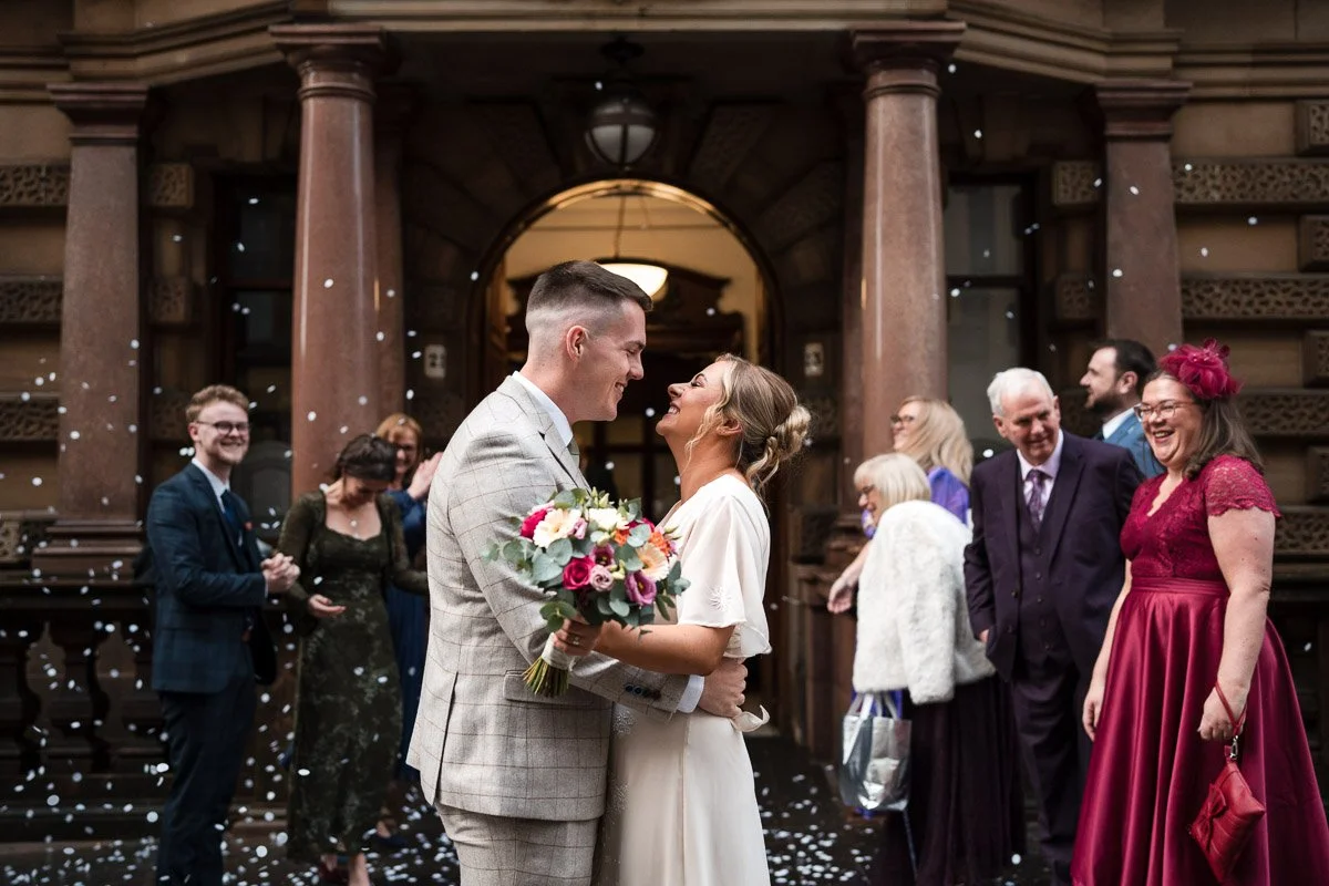 A bride and groom share a kiss in front of family and friends at a wedding reception, with confetti falling around them at Montrose Street, Glasgow