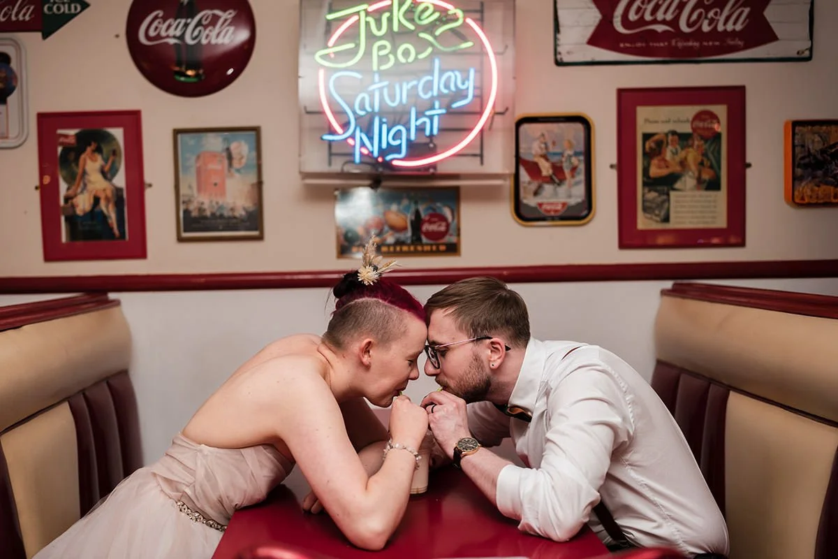A bride and groom sitting in a booth at a restaurant, sharing a milkshake after their Glasgow city elopement. The woman has red hair styled in a bun and is wearing a strapless beige dress. 