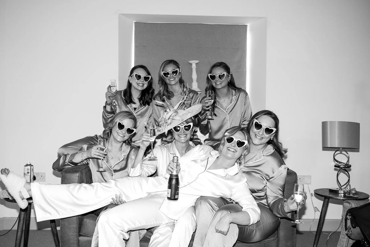 A group of women during bridal prep wearing matching satin pajamas and heart-shaped sunglasses, celebrating with champagne in a living room.