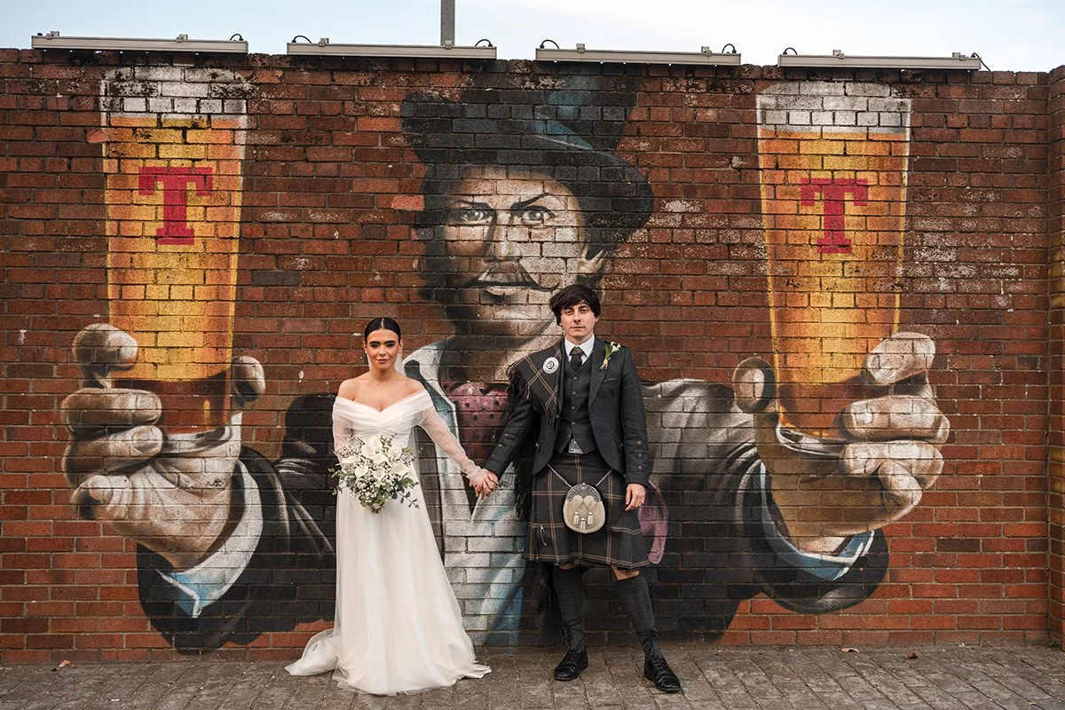 A bride and groom holding hands in front of a large mural of a man holding two pints of beer on a brick wall.