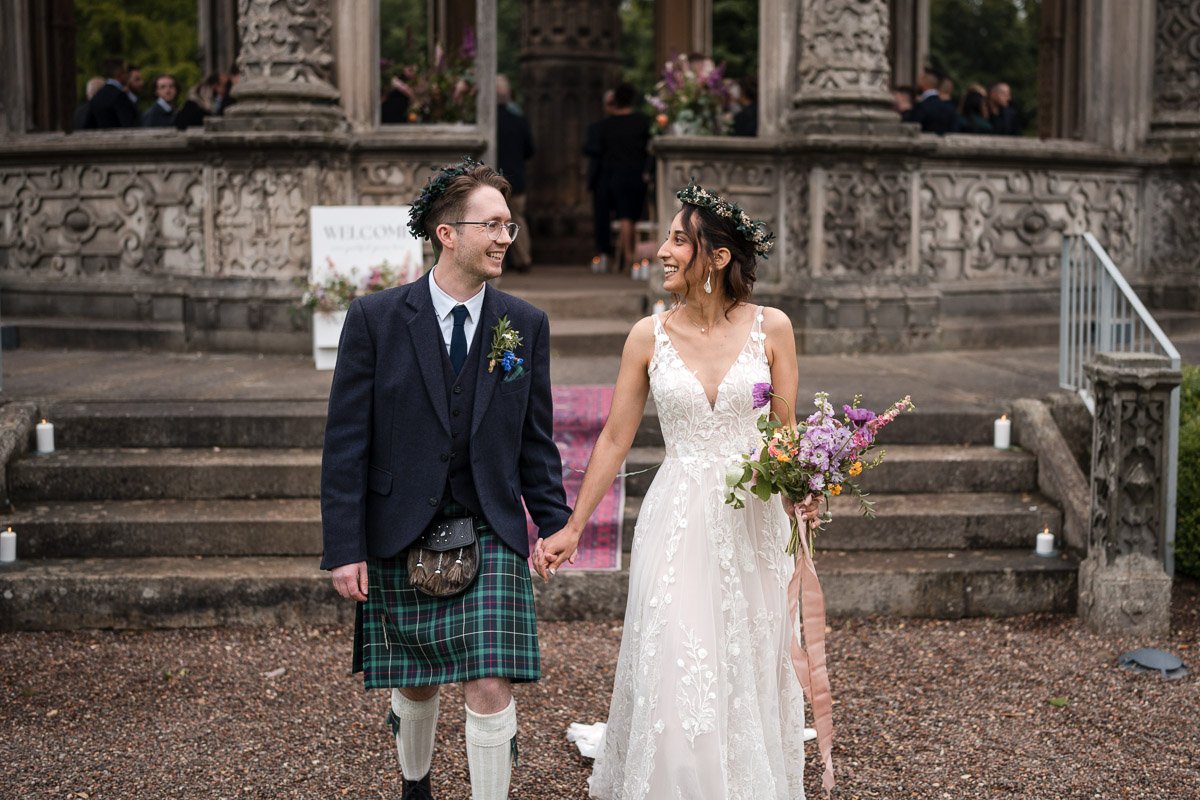 A bride and groom holding hands, smiling at each other in front of a historic stone building with stairs, candles, and floral decorations for a wedding at Restoration Yard Dalkeith