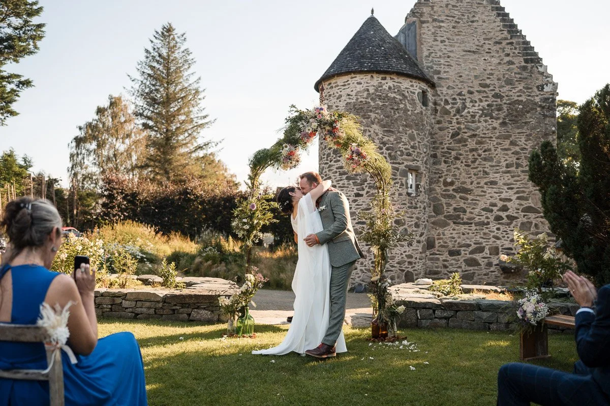 A newlywed couple sharing a kiss at an outdoor wedding ceremony under a floral arch, with guests taking photos, in front of an old stone building, during sunset at Kilmartin Castle