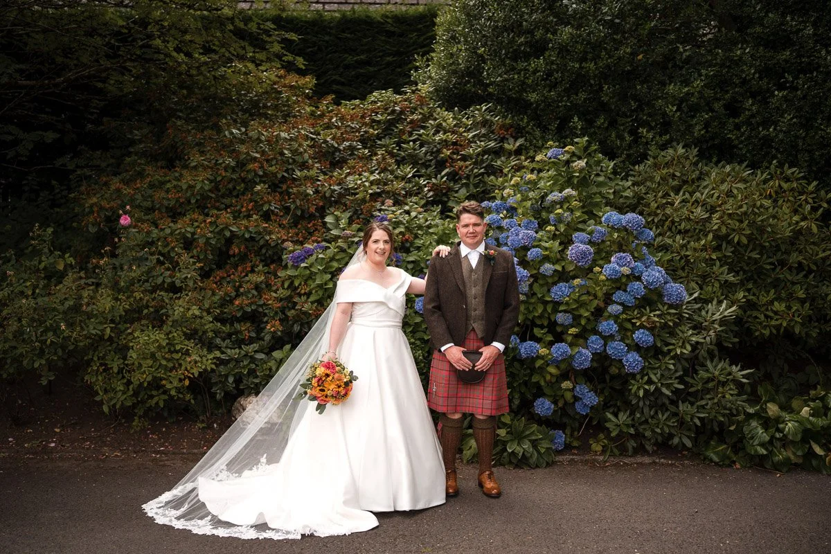 A bride in a white wedding dress holding a bouquet of flowers and a groom in traditional Scottish attire, including a kilt, standing outdoors in front of lush green bushes and blue hydrangeas.