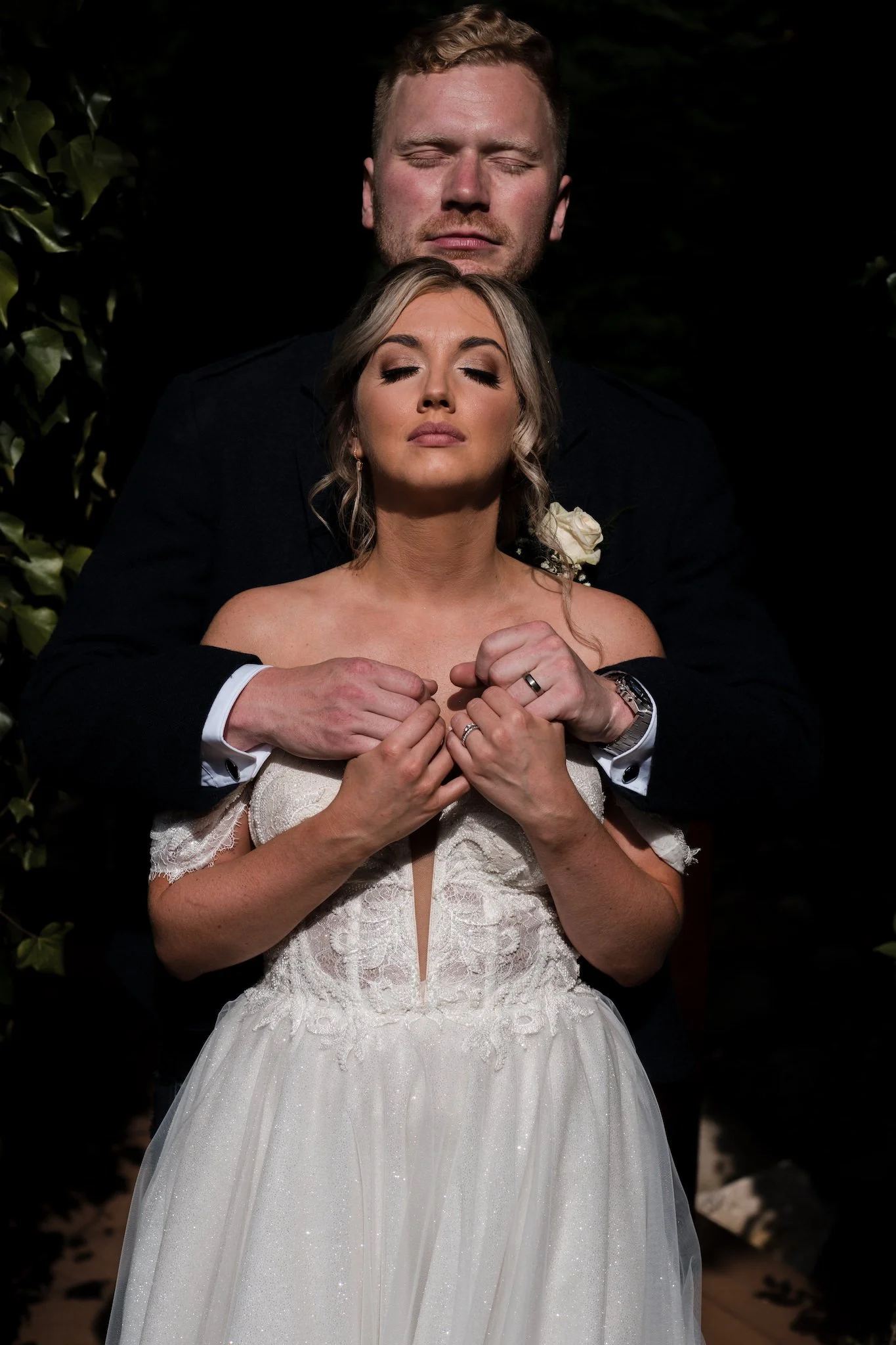 A bride and groom stand close together, with the groom embracing the bride from behind. Both have their eyes closed and appear to be in a serene, emotional moment. The bride is wearing a white wedding dress with lace details, and the groom is dressed