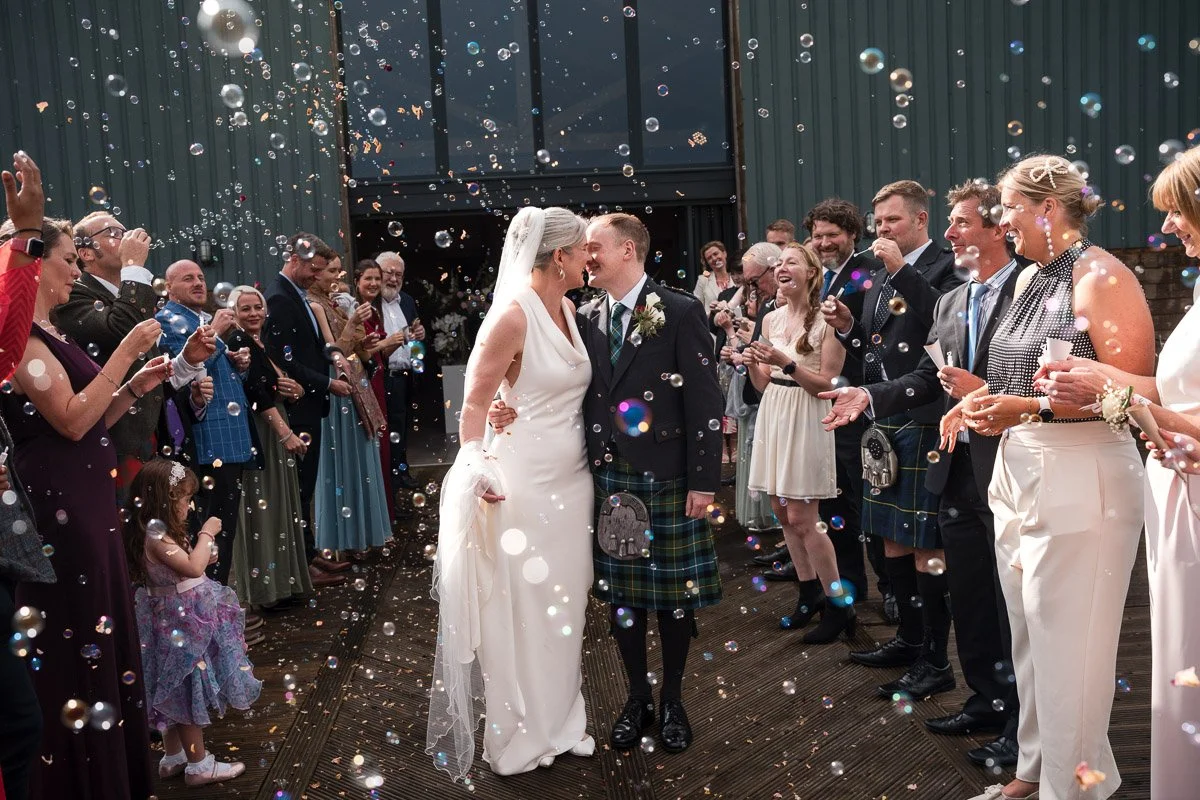 Bride and groom sharing a kiss while surrounded by wedding guests throwing confetti and bubbles at Roodlea Barn