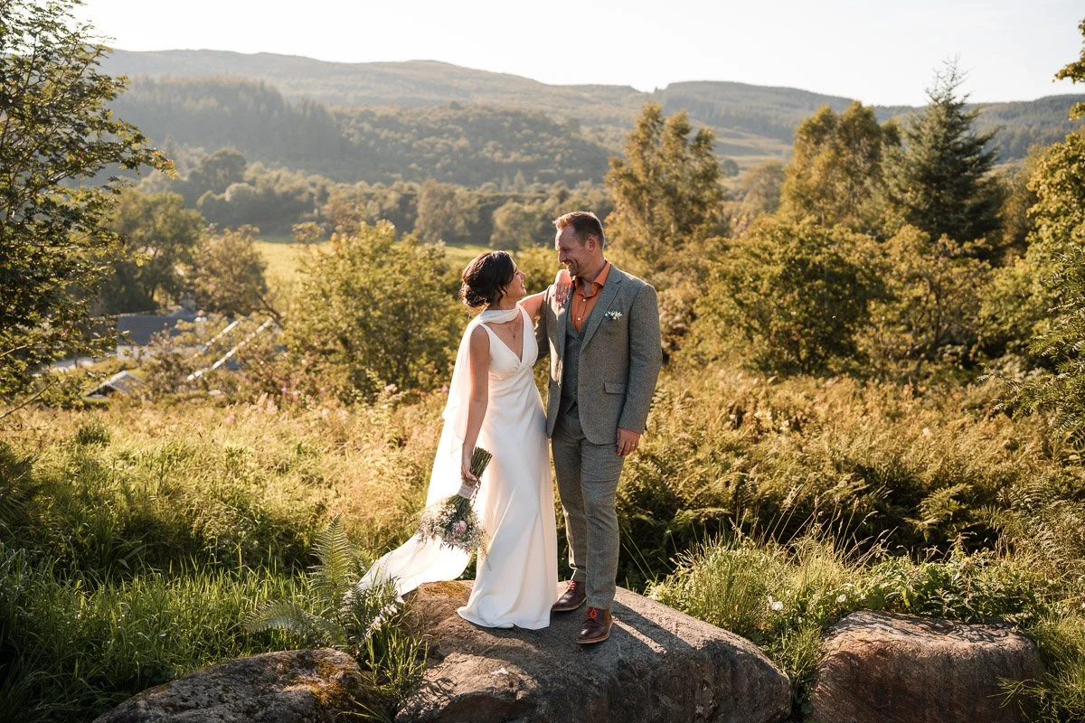 A bride and groom smiling at each other on a large rock in a scenic outdoor landscape with mountains, trees, and sunlight at Kilmartin Castle