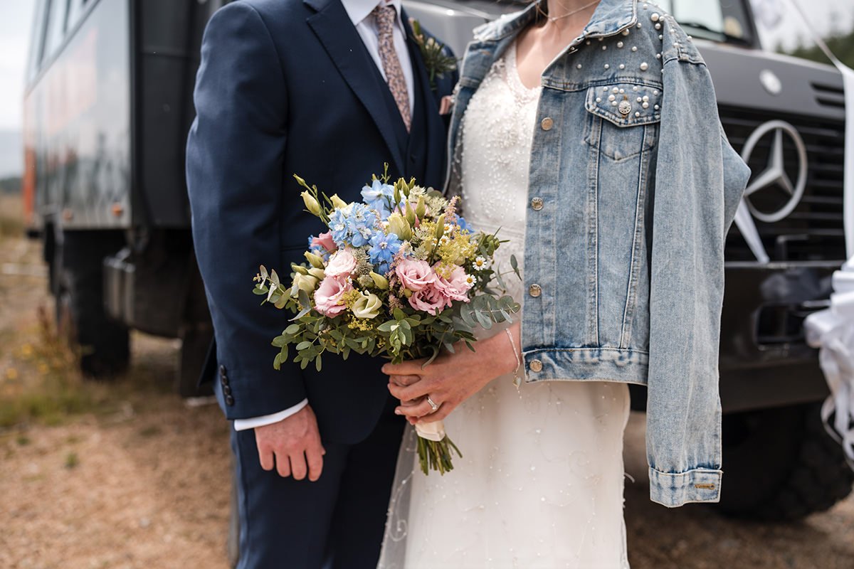 Bride holding a bouquet during an Isle of Arran elopement, wearing a wedding dress with a denim jacket beside the groom.