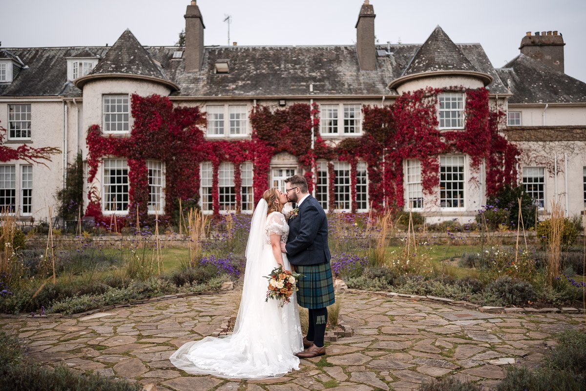 A bride and groom sharing a kiss in front of a large, historic mansion with ivy-covered windows and turrets on a cloudy day at Rufflets, St. Andrews
