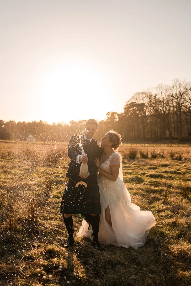A bride and groom celebrating outdoors at sunset, with the groom spraying champagne and both smiling. The bride is in a white wedding dress, and the groom is wearing a kilt. They are standing in a field at sunset at Enterkine House Hotel