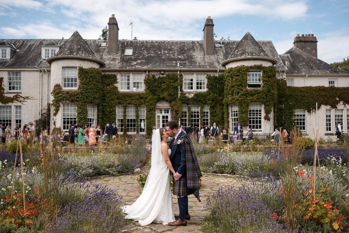 A bride and groom kissing in front of a large historic house with ivy-covered walls, surrounded by a colorful garden, during a wedding celebration at Rufflets, St. Andrews