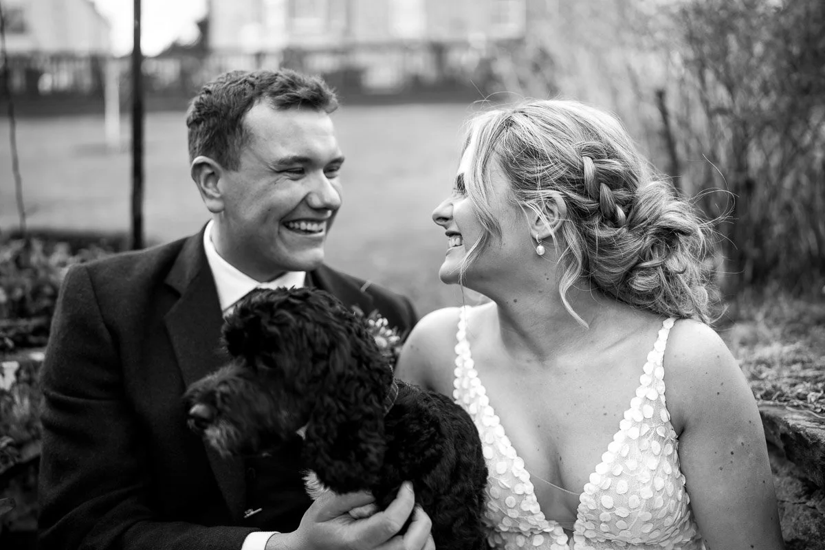 A couple in wedding attire smiling at each other with a black puppy in the groom's hands, outdoors in black and white at Loch Ness House Hotel