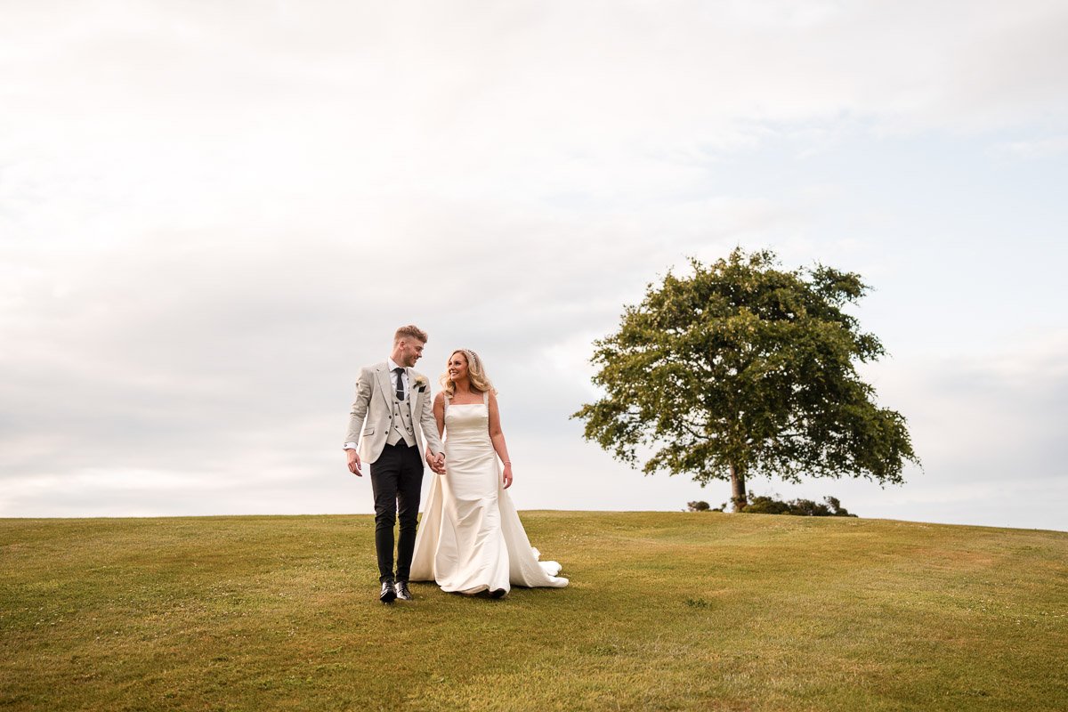 A bride and groom walking hand in hand on a grassy field with a large tree in the background and a cloudy sky at a wedding at Fairmont, St. Andrews