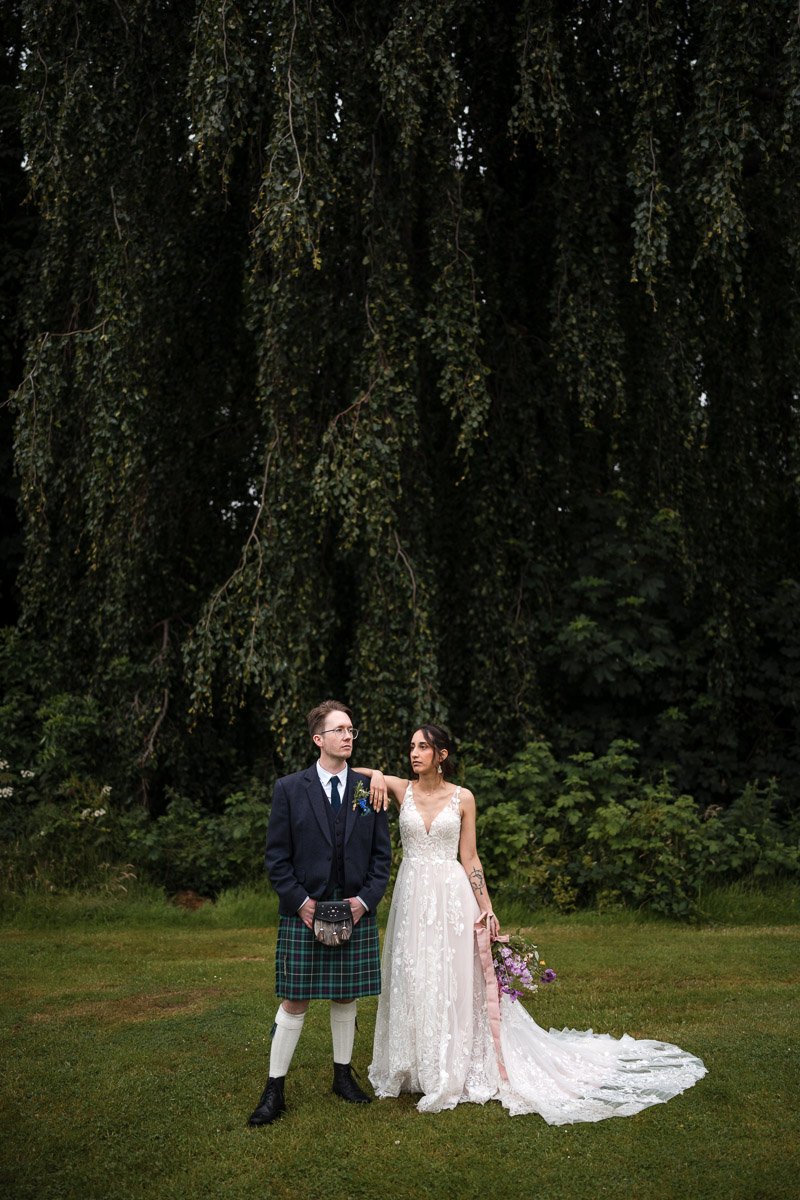 A bride and groom standing on grass in a wooded outdoor setting at Restoration Yard, Dalkeith. The groom is wearing traditional Scottish attire. The bride is in a white lace wedding dress holding a bouquet of purple flowers