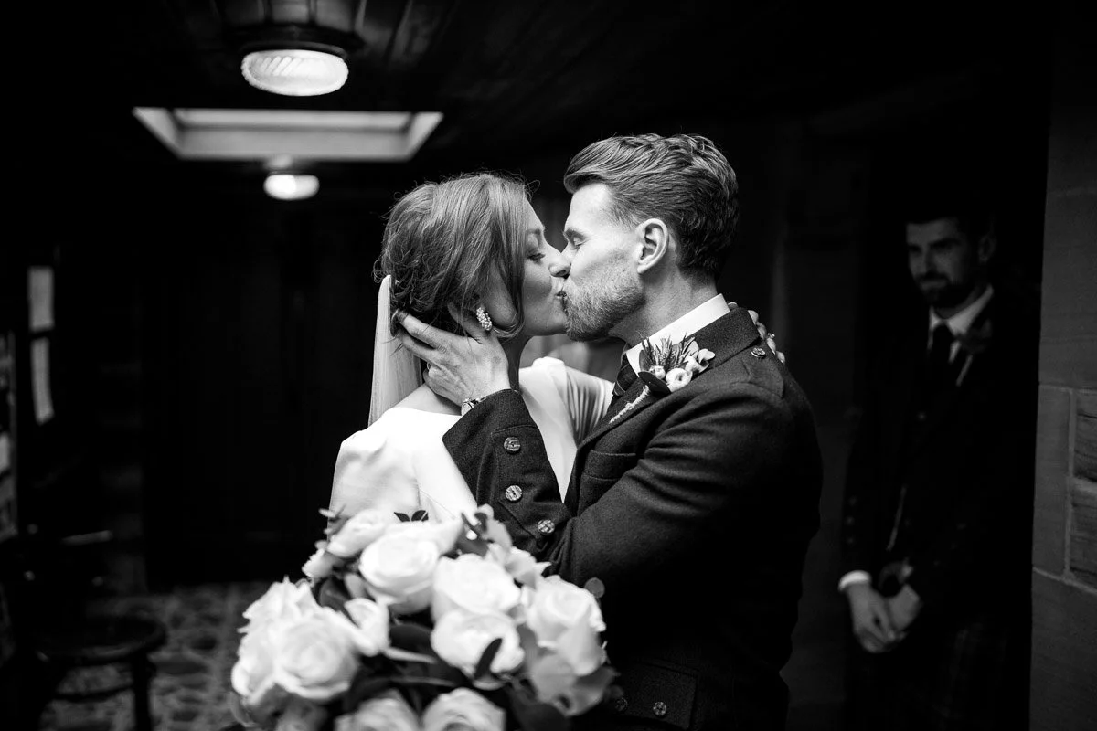 A black and white photo of a bride and groom kissing, holding each other closely. The bride wears wedding attire with a veil and earrings, while the groom wears a suit with a boutonniere. A bouquet of roses is visible in the foreground. A man in a da