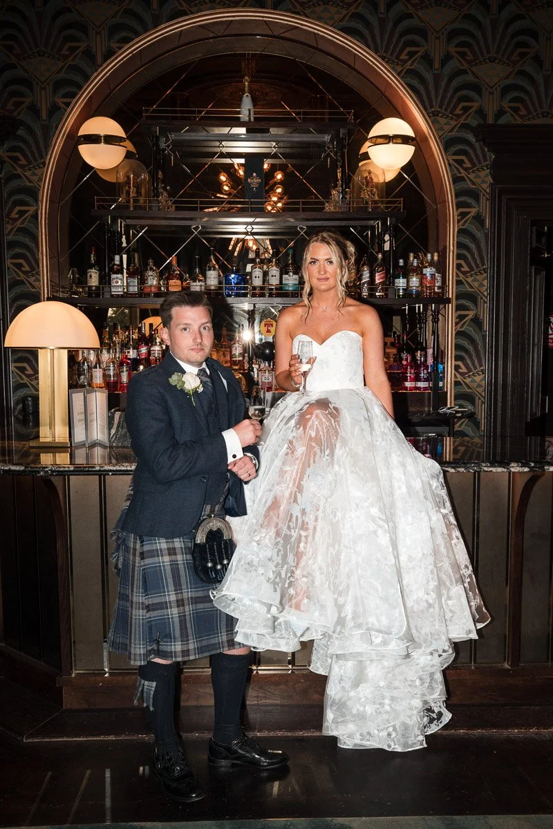 A bride and groom at a bar in Cornhill Castle, with the bride sitting on the bar counter holding a glass, dressed in a white wedding gown, and the groom standing next to her in traditional Scottish attire, holding a glass.