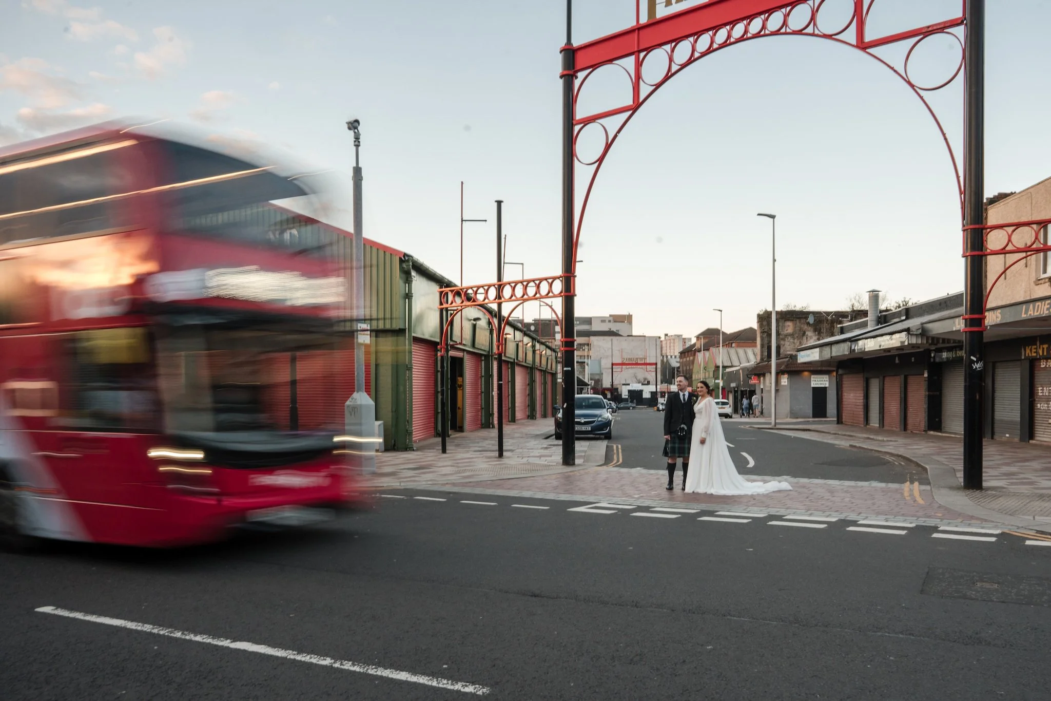 A city street scene with a blurred red double-decker bus passing by, while a bride and groom dressed in wedding attire stands on the corner under a large red metal archway at the Barras Glasgow