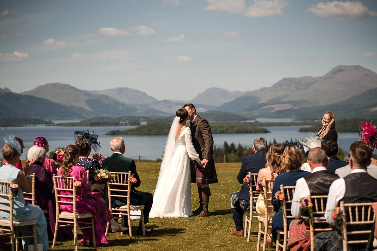 A wedding ceremony taking place outdoors near a loch with mountains in the background at Ardoch, Loch Lomond. The bride and groom are kissing, surrounded by seated guests.