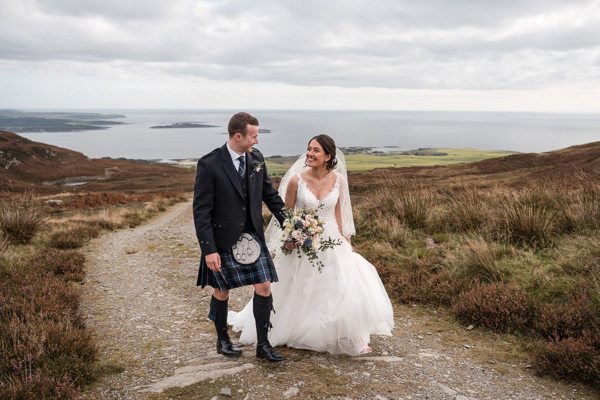 A bride and groom holding hands, walking on a dirt path through a mountainous landscape with a lake in the background, during their wedding day at GG's Yard