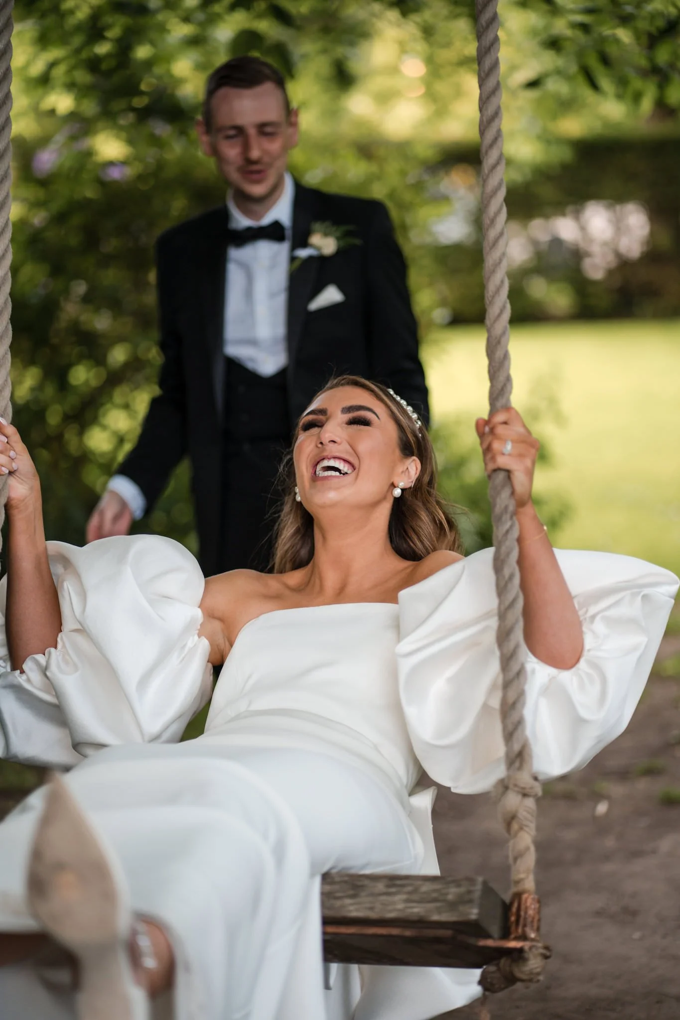 A bride in a white wedding dress swinging on a wooden swing, smiling and laughing, with a groom in a tuxedo standing behind her, outdoors with greenery and trees in the background at Balbirnie House Hotel Wedding