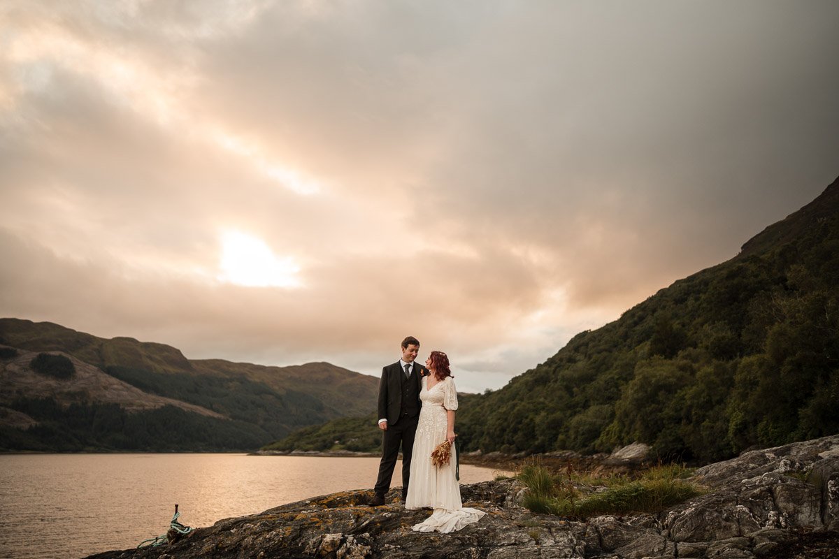 A bride and groom standing on a rocky shoreline by a lake with hills and cloudy sky in the background, during sunset at the elopement in Dunoon