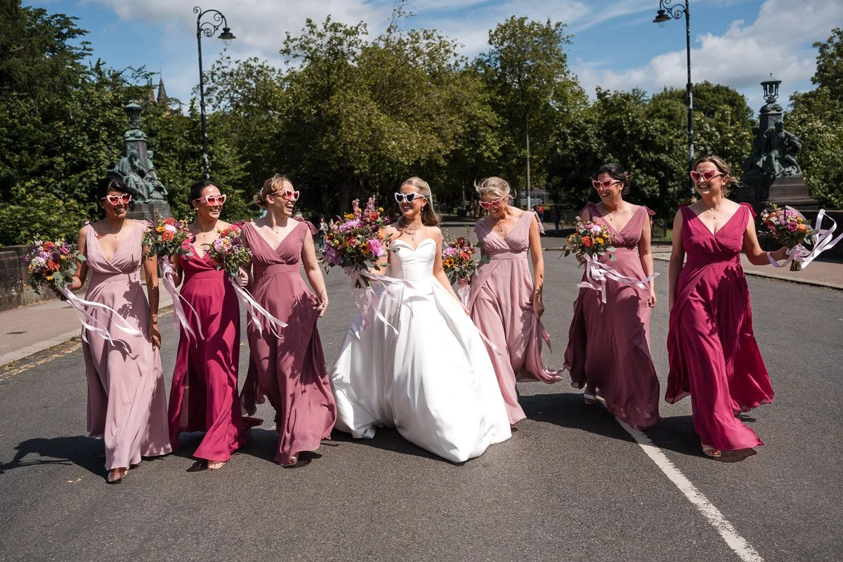 A bride in a white wedding dress walking with six bridesmaids in pink and purple dresses, all holding bouquets and wearing sunglasses, on a city street with trees and lampposts in the background in Glasgow West End