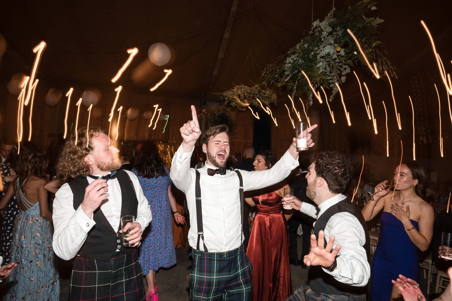 People celebrating at a wedding reception with glasses of drinks, wearing formal and kilts, under string lights and hanging lanterns.