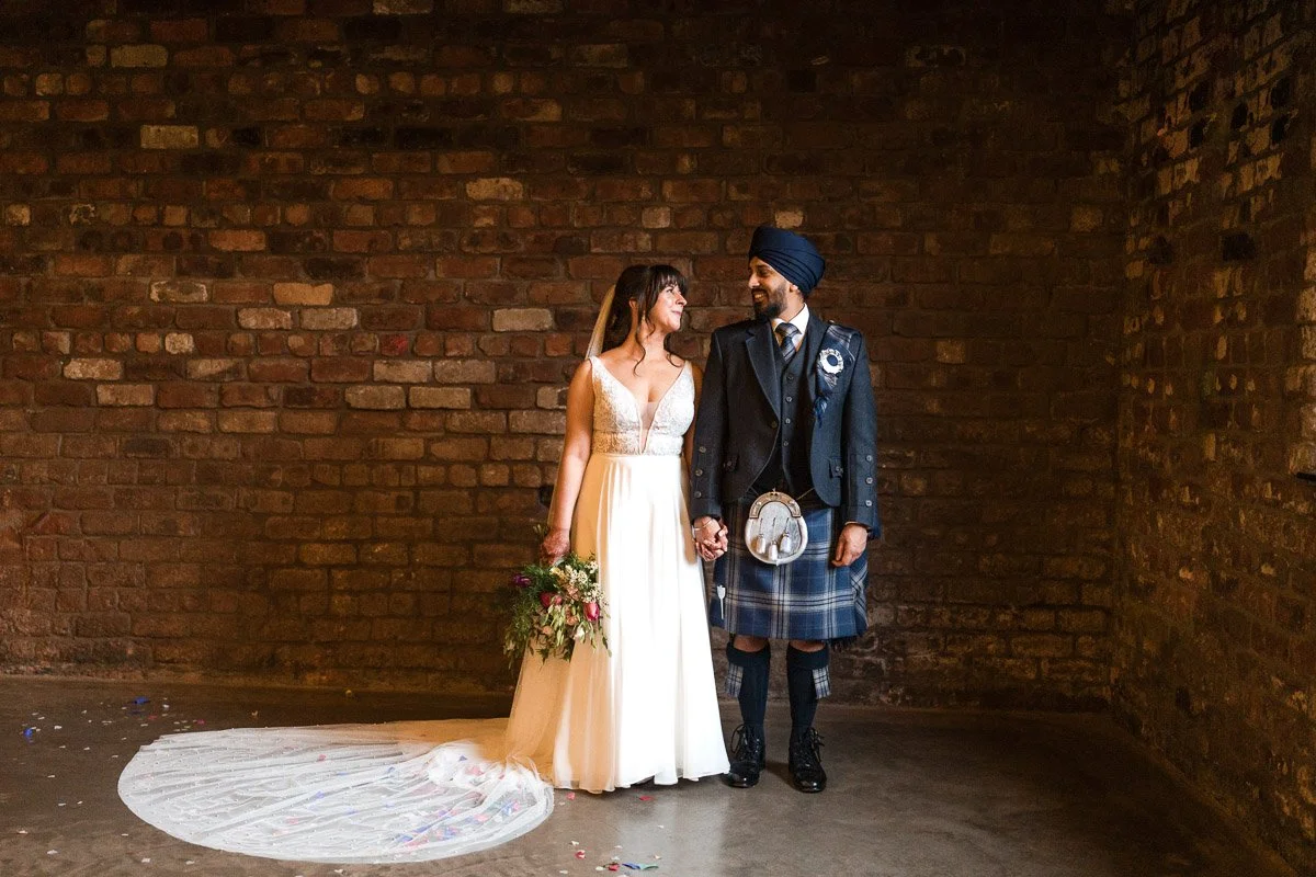 A bride and groom holding hands and smiling at each other against a brick wall on their wedding day. The bride is wearing a white wedding dress and holding a bouquet, and the groom is dressed in traditional Scottish attire and turban at Engine Works