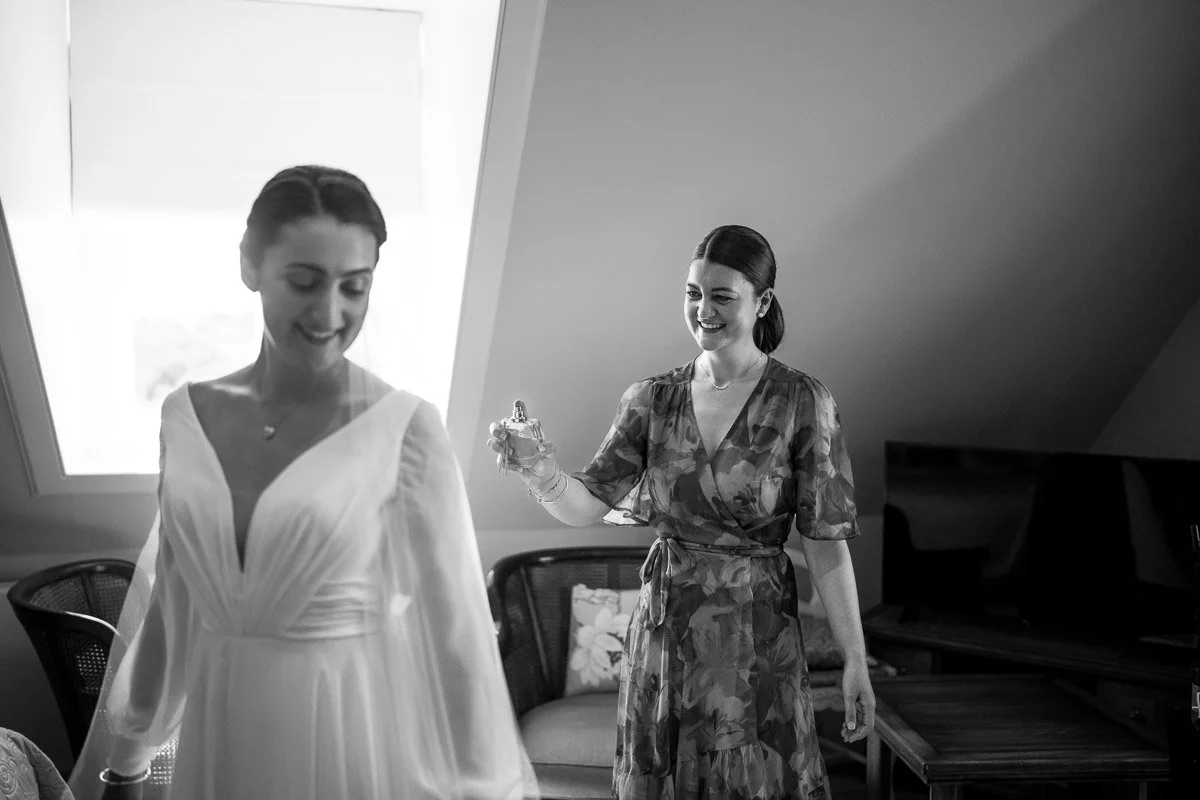 Two women, one in a wedding dress and one in a floral dress, smiling and preparing in a room with a slanted ceiling and a window at Ardoch, Loch Lomond