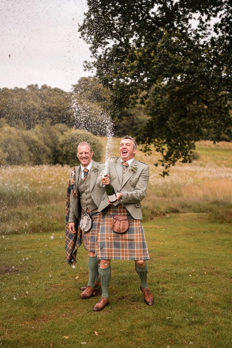 Two men in traditional Scottish kilts and suits celebrating outdoors, one opening a champagne bottle with a spray of champagne around them, in a grassy area with trees in the background at Eden Leisure Village