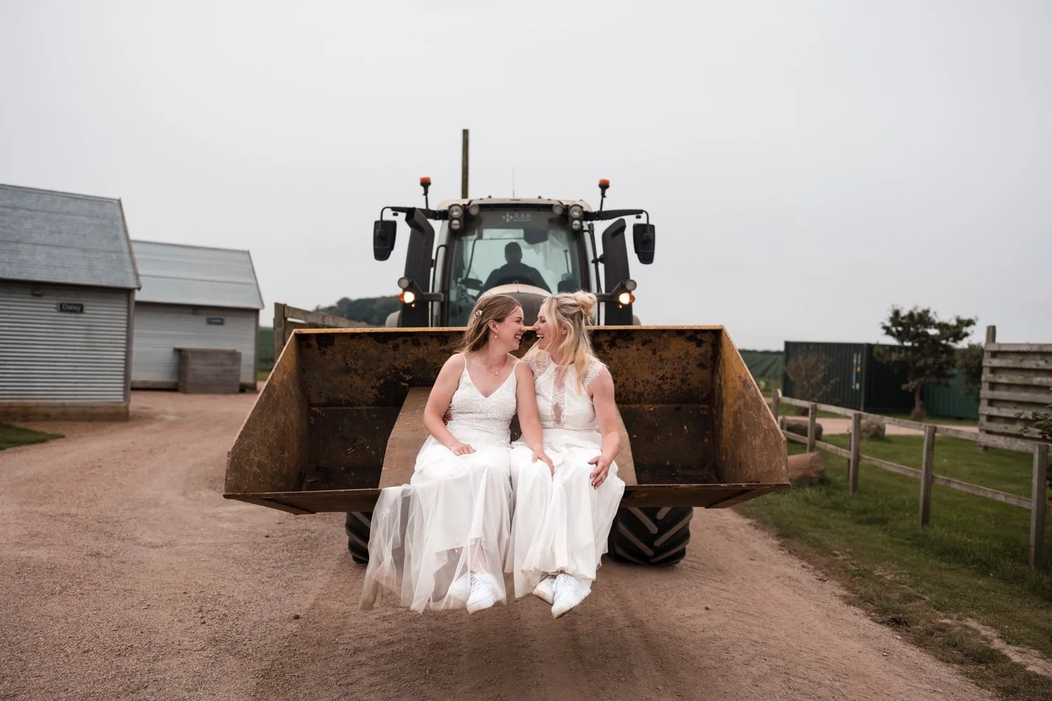 Two brides in white dresses sitting on a tractor bucket, smiling and touching noses, with a tractor behind them in a farm setting on Cow Shed, Crail.