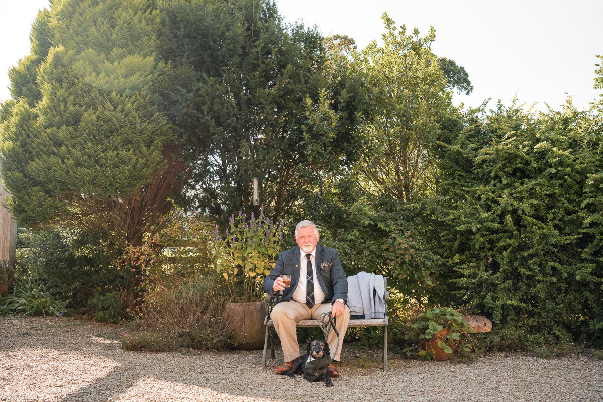 An elderly man with white hair and a beard sitting on a garden bench, holding a glass of dark beverage, with a black dog sitting in front of him. The man is dressed in a shirt, tie, and jacket at Kilmartin Castle during a wedding