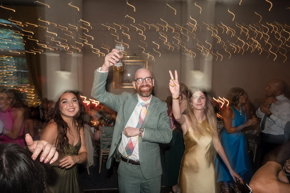 People celebrating at a party, with a man in a suit raising a drink and making a peace sign, surrounded by smiling women and other guests, with festive string lights overhead at Balbirnie House