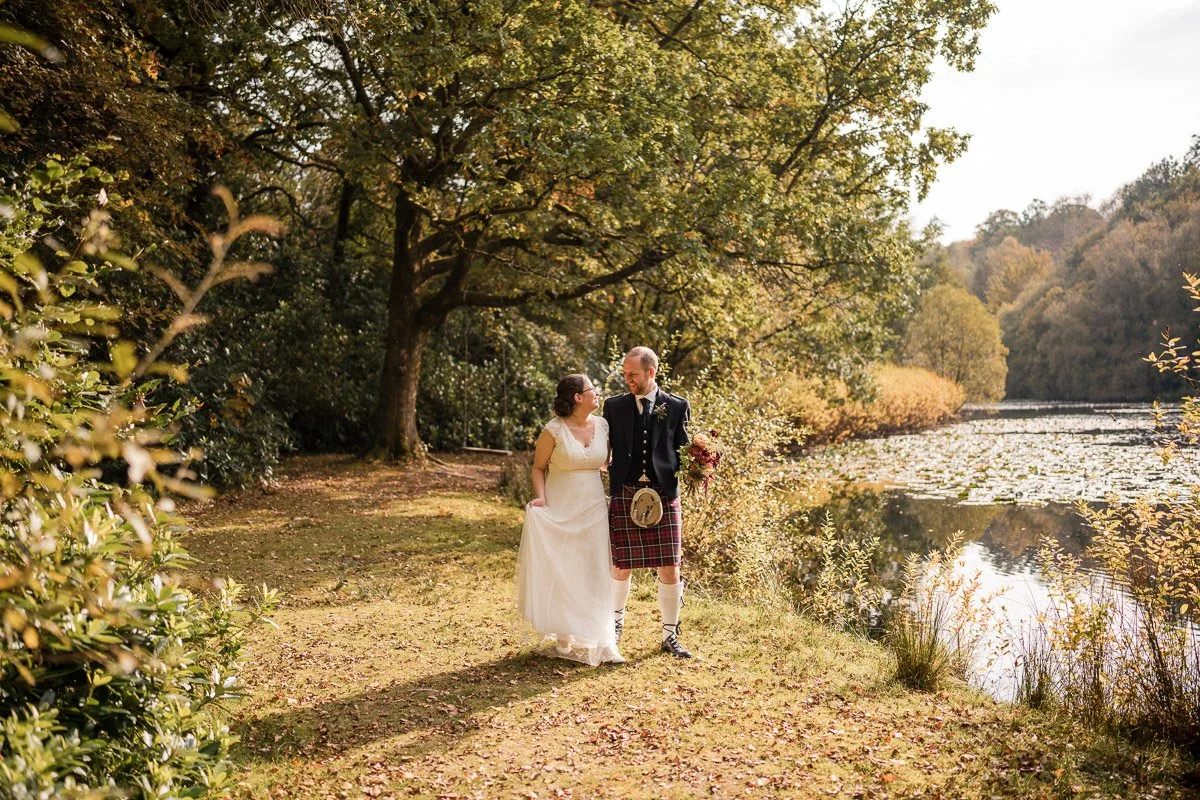 A bride and groom walk together along a wooded lakeside trail on their wedding day, surrounded by trees with autumn foliage, water lilies on the lake, and warm sunlight at their wedding at Auchen Castle