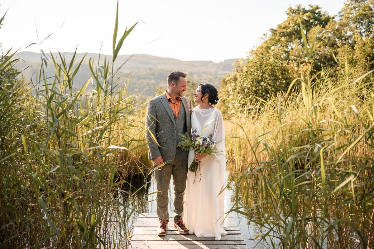 A couple dressed in wedding attire standing on a wooden bridge in a field of tall grass, smiling and looking at each other during sunset at the Kilmartin Castle elopement