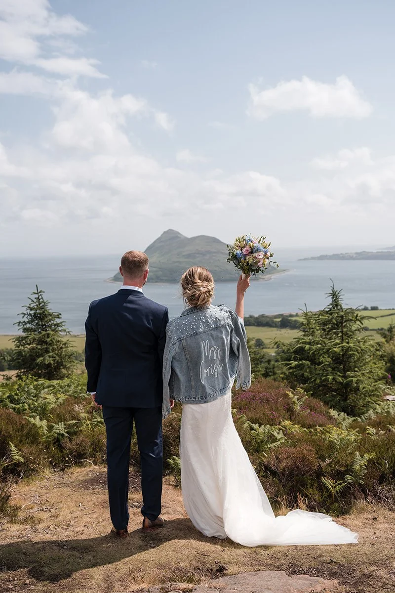 A bride and groom stand together outdoors. The bride is holding a bouquet of flowers and is wearing a denim jacket. They are overlooking a scenic landscape with a lake, small islands, and a mountain in the distance on Arran