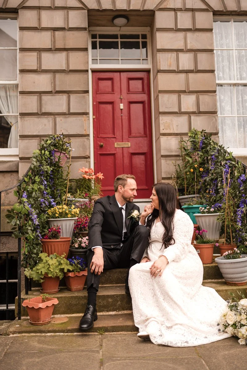 A bride and groom dressed in wedding attire sitting on steps in front of a house with potted flowers and plants around them after their Edinburgh elopement.