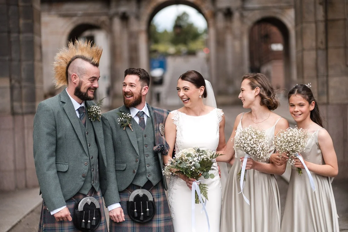 A wedding party standing outdoors under a stone arch, smiling and laughing, with three men in kilts and a bride and two young girls in bridesmaid dresses holding bouquets at their intimate wedding in Glasgow City Centre