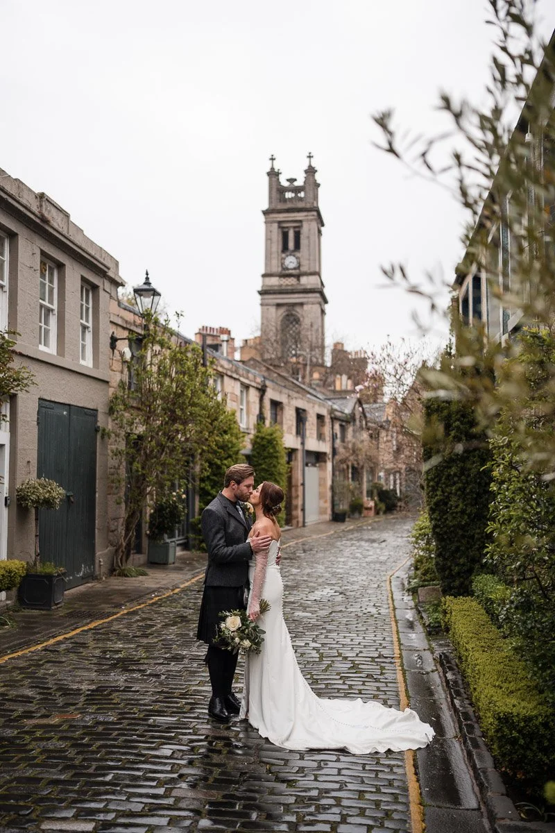 A bride and groom standing close on a cobblestone street in Circus Lane, Edinburgh with historic buildings and a clock tower in the background, after a city elopement in Edinburgh