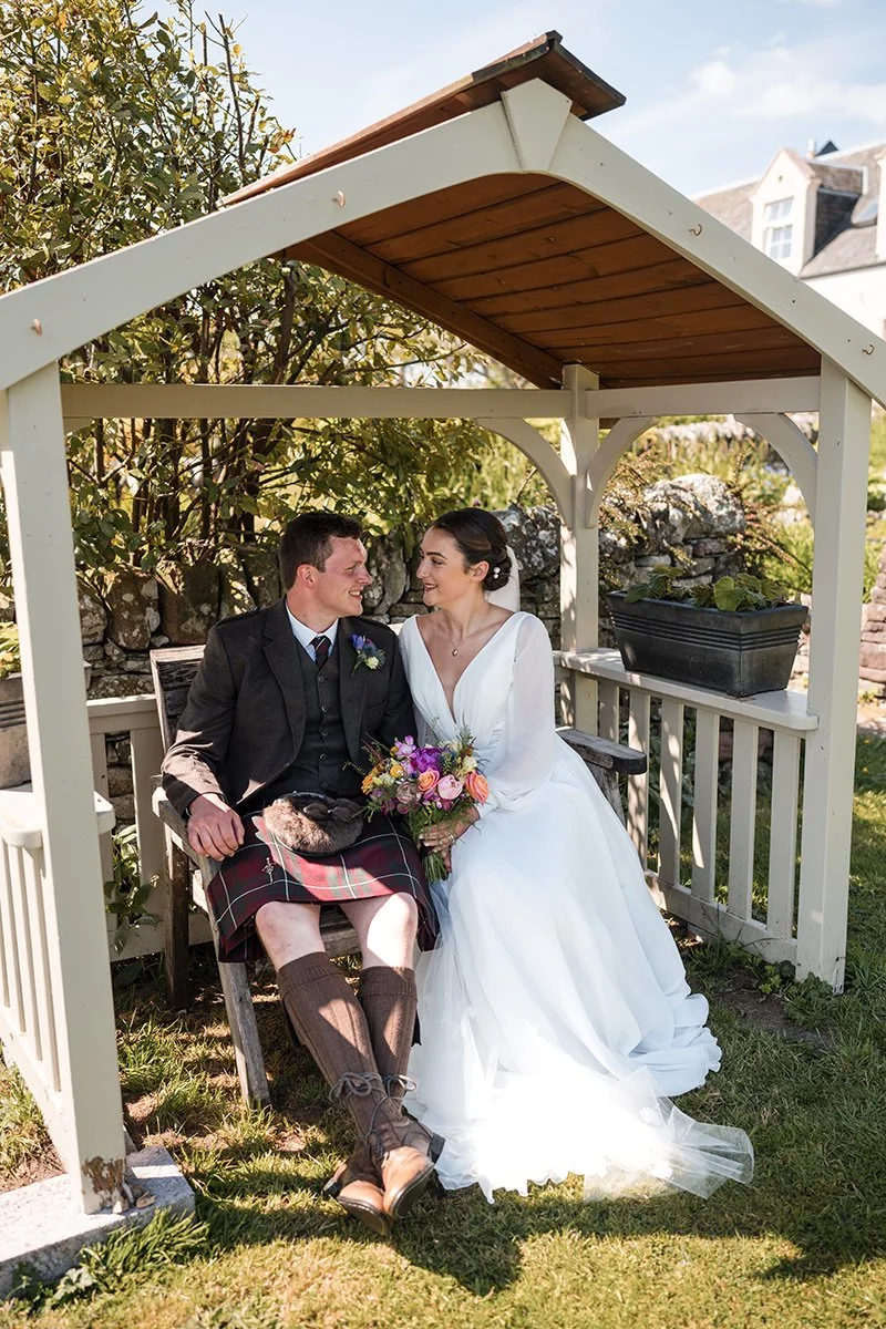 A bride and groom sitting together inside a small garden gazebo at Ardoch, Loch Lomond. The groom is wearing traditional Scottish kilt and jacket, holding a tartan plaid, and the bride is wearing a white wedding gown holding a colourful bouquet of fl