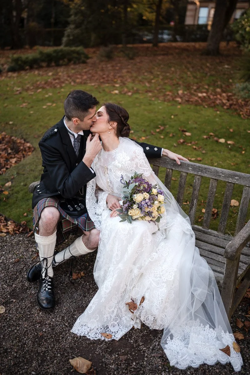 A newlywed couple sharing a kiss on a park bench. The groom is dressed in a black jacket, kilt, and sporran, and the bride wears a white lace wedding gown holding a bouquet of flowers with purple and cream colors. Fall leaves are on the ground.