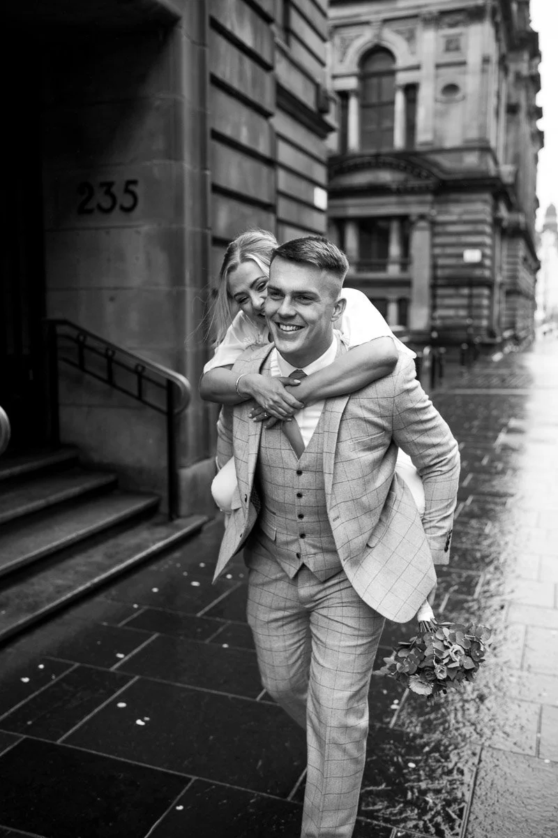 A happy groom in a checkered suit giving a piggyback ride to his bride, holding a bouquet of flowers, on a city street in black and white after their Glasgow City Centre elopement